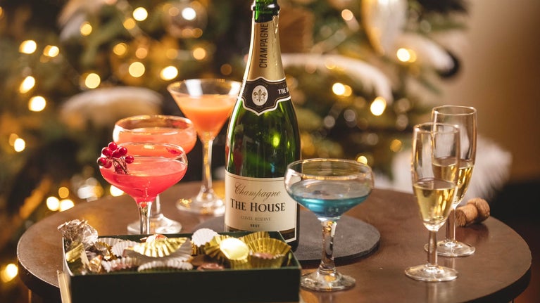 Festive scene with a bottle of champagne, colorful cocktails, and chocolates on a table. A Christmas tree with warm lights is glowing in the background.